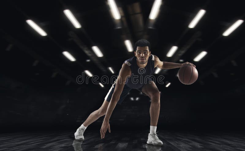 Young African sportsman, basketball player training in gym, idoors isolated on dark background. Concept of sport, game