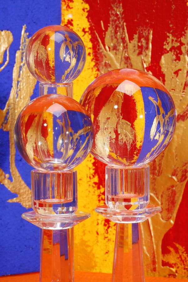 Three balls of crystal glass of different sizes, two of them on candlesticks, one on a tube of glass. Background is of vivid blue, red and gold, bottom in orange. All these colours are reflected in the glass. Three balls of crystal glass of different sizes, two of them on candlesticks, one on a tube of glass. Background is of vivid blue, red and gold, bottom in orange. All these colours are reflected in the glass.