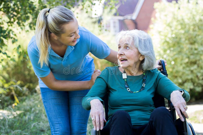 Carer Pushes Senior Woman In Wheelchair In Garden. Carer Pushes Senior Woman In Wheelchair In Garden