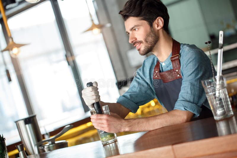 Smiling male bartender wiping a glass and looking away. Barista in