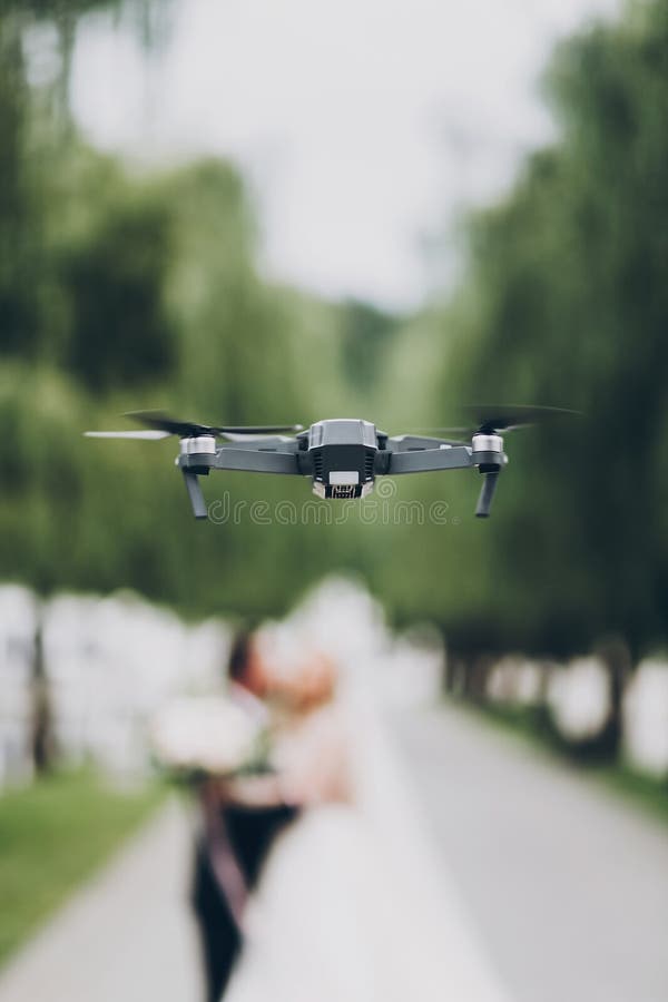 Drone flying in green part. Drone photo and footage of wedding day concept. Black drone on background of blurred wedding couple in green park. Drone flying in green part. Drone photo and footage of wedding day concept. Black drone on background of blurred wedding couple in green park