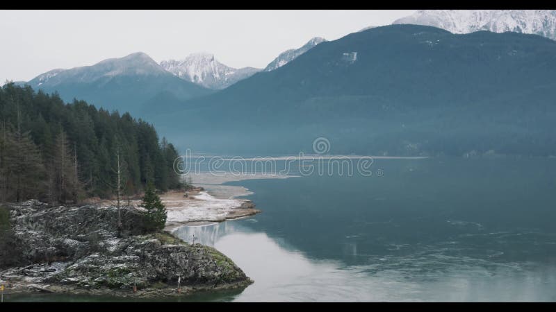 Drone scenery of a lake with rocky shore with dense trees and snowy mountain in the background stock video footage