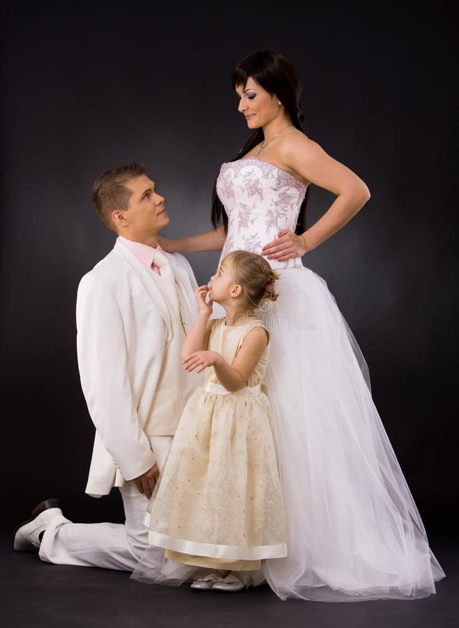 Portrait of wedding couple and little girl bridesmaid. Bride wearing romantic white wedding dress, groom kneealing if front of her. Portrait of wedding couple and little girl bridesmaid. Bride wearing romantic white wedding dress, groom kneealing if front of her.