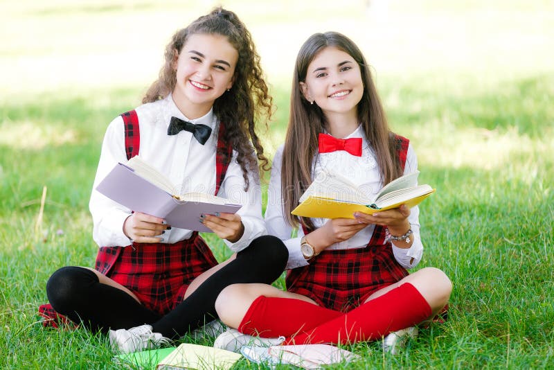 Two pretty schoolgirls in school uniforms sit with books outdoors in the park. Schoolgirls or students are taught lessons in nature. Two pretty schoolgirls in school uniforms sit with books outdoors in the park. Schoolgirls or students are taught lessons in nature.