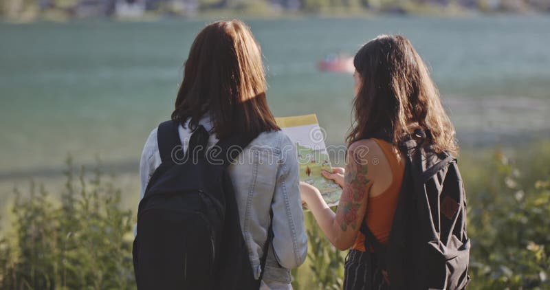 Duas garotas turistas olham para o mapa na costa do rio. belas morenas com mochila à procura de um guia no ensolarado