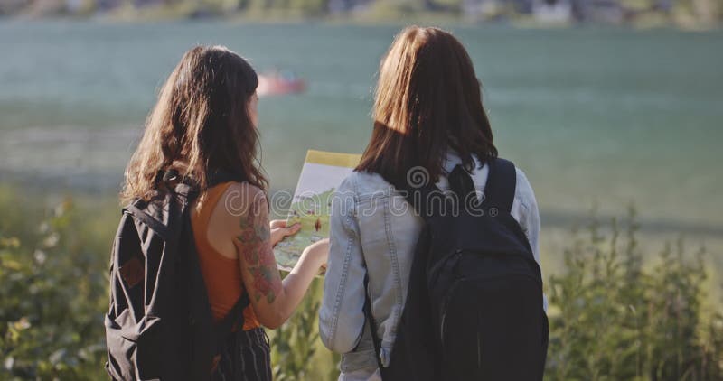 Duas garotas turistas olham para o mapa na costa do rio. belas morenas com mochila à procura de um guia no ensolarado