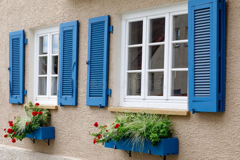 Duas janelas com quadros brancos de madeira, obturadores azuis e caixas decorativas de flores Imagem do decor da moda, conforto