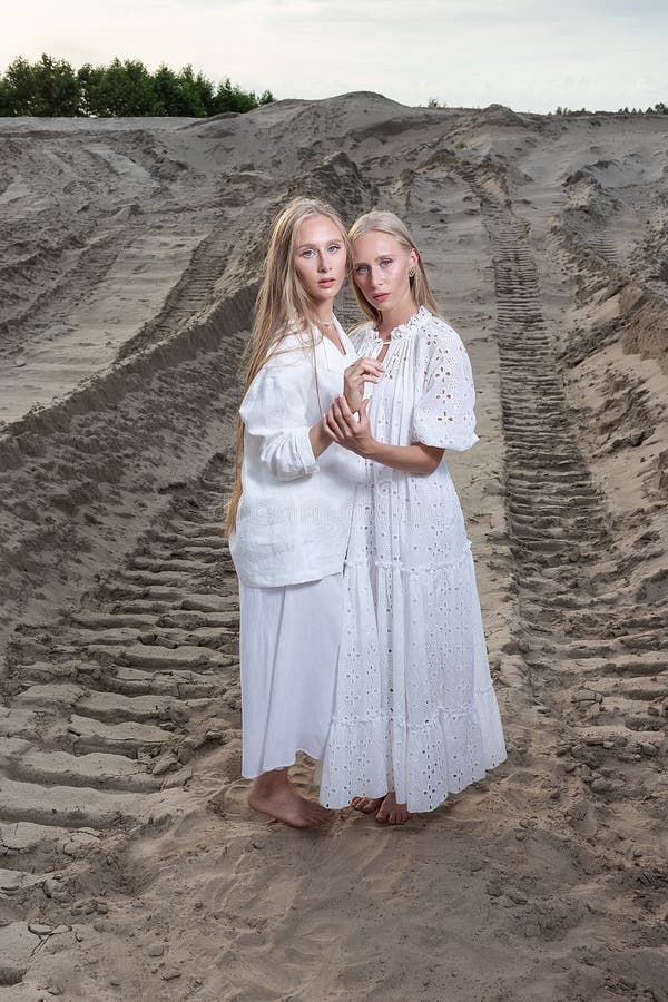 Young pretty twins with long blond hair hold hands at sand quarry in elegant white dress, skirt, jacket. stylish fashion photoshoot, summer photosession. identical sisters spend time together outdoors. Young pretty twins with long blond hair hold hands at sand quarry in elegant white dress, skirt, jacket. stylish fashion photoshoot, summer photosession. identical sisters spend time together outdoors