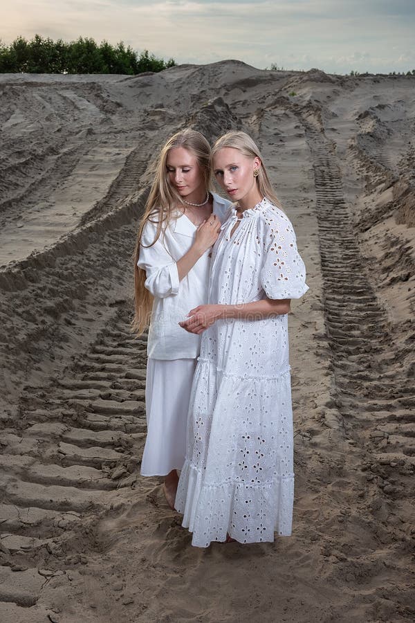 Young blond pretty twins posing at sand quarry in elegant white dress, skirt, jacket. stylish fashion photoshoot with flashlight, summer photosession. identical sisters spend time together outdoors. Young blond pretty twins posing at sand quarry in elegant white dress, skirt, jacket. stylish fashion photoshoot with flashlight, summer photosession. identical sisters spend time together outdoors