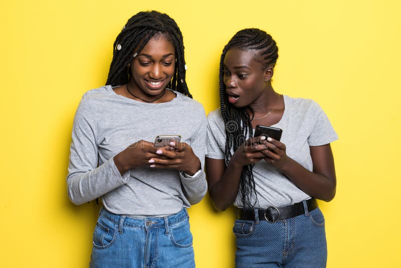 Duas jovens mulheres africanas que espiam-se quando ambos que usam os telefones celulares isolados sobre o fundo amarelo fotografia de stock royalty free Duas jovens mulheres africanas que espiam-se quando ambos que usam os telefones celulares isolados sobre o fundo amarelo fotografia de stock royalty free