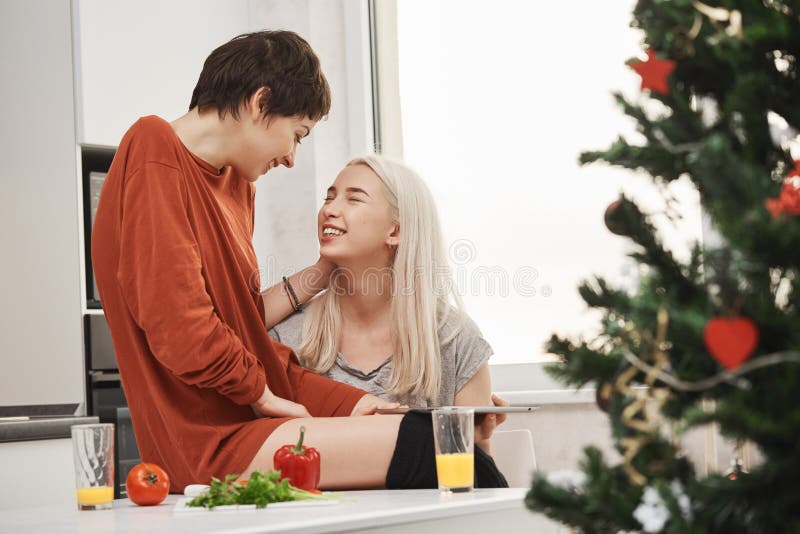 Duas meninas bonitos que sentam-se na cozinha ao falar e ao rir durante o café da manhã perto da árvore de Natal Manhã feliz típi imagem de stock royalty free Duas meninas bonitos que sentam-se na cozinha ao falar e ao rir durante o café da manhã perto da árvore de Natal Manhã feliz típi imagem de stock royalty free