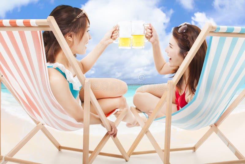Duas meninas da luz do sol que guardam elogios da cerveja em uma cadeira de praia imagens de stock Duas meninas da luz do sol que guardam elogios da cerveja em uma cadeira de praia imagens de stock