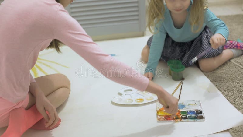 Duas meninas pintam com tintas aquáticas e pincéis em um grande papel de desenho no chão criatividade articular infantil.