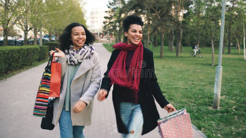Duas mulheres atrativas da raça misturada que dançam e têm o divertimento ao andar abaixo do parque com sacos de compras Amigos n foto de stock royalty free Duas mulheres atrativas da raça misturada que dançam e têm o divertimento ao andar abaixo do parque com sacos de compras Amigos n foto de stock royalty free