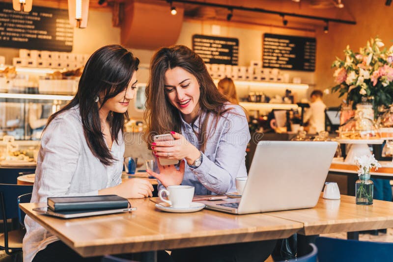 Duas mulheres felizes novas estão sentando-se no café na tabela na frente do portátil, usando o smartphone e o riso