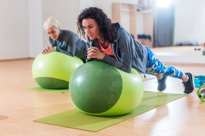 Duas mulheres positivas que fazem a prancha exercitam o encontro na bola do equilíbrio no gym