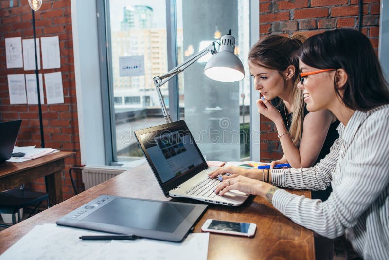 Duas mulheres que trabalham no Web site novo projetam a escolha de imagens usando o portátil que surfa o Internet imagem de stock royalty free Duas mulheres que trabalham no Web site novo projetam a escolha de imagens usando o portátil que surfa o Internet imagem de stock royalty free