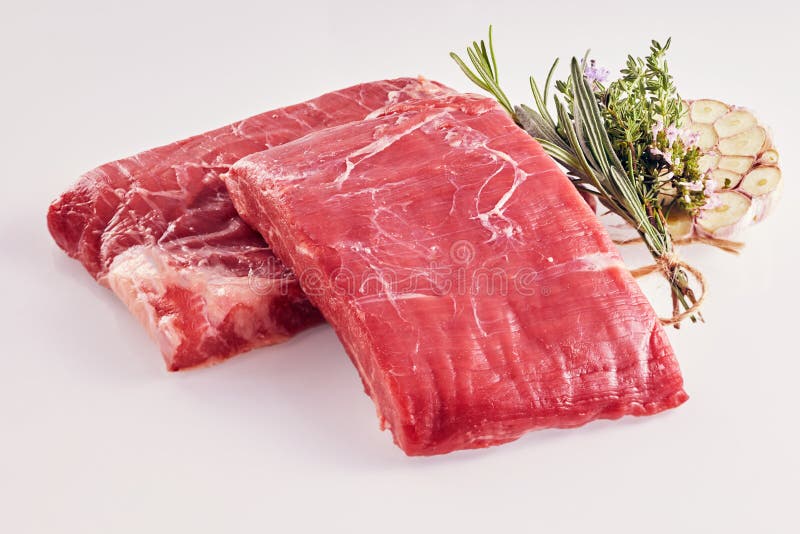 Two rectangular portions of raw lean flank steak with fresh garlic cloves and a bouquet garni with fresh herbs including rosemary and thyme. Two rectangular portions of raw lean flank steak with fresh garlic cloves and a bouquet garni with fresh herbs including rosemary and thyme
