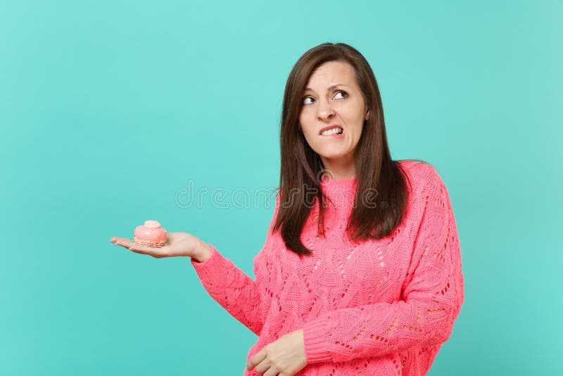 Dubitando giovane donna in maglione rosa tricottato che cerca, tenente dolce disponibile e mordente le labbra isolate su turchese fotografia stock Dubitando giovane donna in maglione rosa tricottato che cerca, tenente dolce disponibile e mordente le labbra isolate su turchese fotografia stock