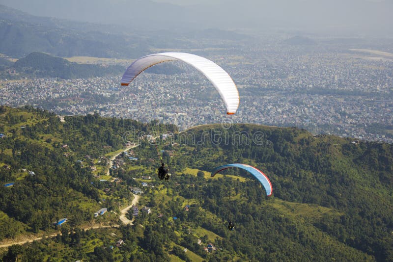 Due alianti su un paracadute bianco e blu sorvolano le montagne verdi una città nella valle immagini stock libere da diritti Due alianti su un paracadute bianco e blu sorvolano le montagne verdi una città nella valle immagini stock libere da diritti