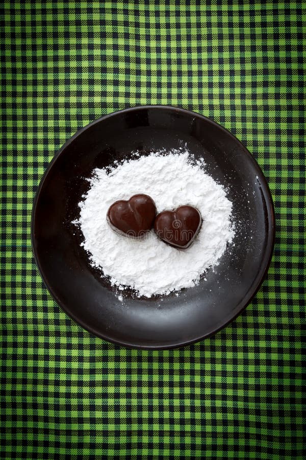 Due caramelle in forma di cuore del cioccolato su un piatto marrone con la polvere dello zucchero contro verde hanno controllato fotografie stock libere da diritti Due caramelle in forma di cuore del cioccolato su un piatto marrone con la polvere dello zucchero contro verde hanno controllato fotografie stock libere da diritti