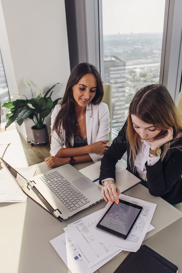 Due colleghi femminili sorridenti che lavorano insieme nell'ufficio che si siede allo scrittorio facendo uso del pc della compres fotografia stock Due colleghi femminili sorridenti che lavorano insieme nell'ufficio che si siede allo scrittorio facendo uso del pc della compres fotografia stock