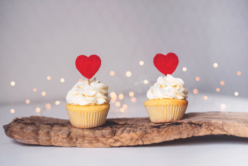 Delicious two white cupcakes with white whipped cream and red heart decoration on top for Valentine`s Day stand on a wooden deck.  With bokeh. Delicious two white cupcakes with white whipped cream and red heart decoration on top for Valentine`s Day stand on a wooden deck.  With bokeh