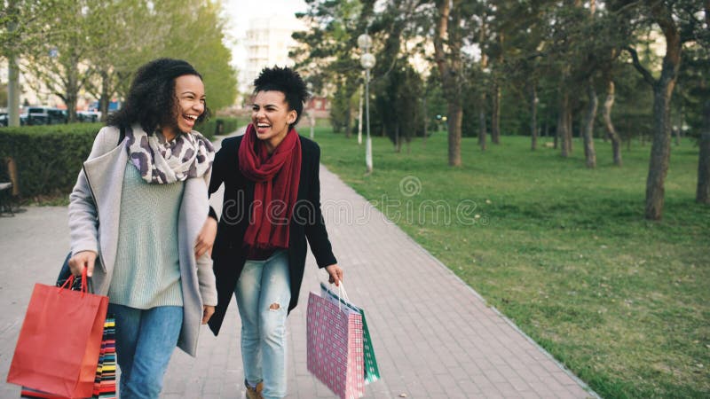 Due donne attraenti della corsa mista che ballano e si divertono mentre camminano giù il parco con i sacchetti della spesa Giovan fotografia stock libera da diritti Due donne attraenti della corsa mista che ballano e si divertono mentre camminano giù il parco con i sacchetti della spesa Giovan fotografia stock libera da diritti