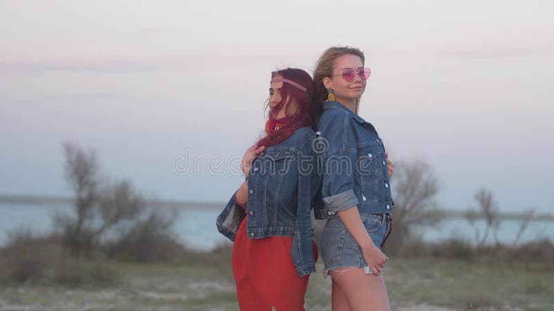 Due giovani donne vestite in rivestimenti del denim per stare indietro per appoggiare nei raggi delicati del tramonto con capelli