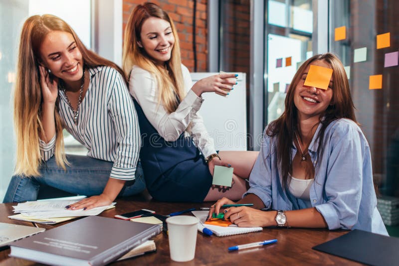 Due ragazze che ridono del loro amico con una nota appiccicosa sul suo fronte Gruppo di studentesse che si rilassano divertendosi fotografia stock Due ragazze che ridono del loro amico con una nota appiccicosa sul suo fronte Gruppo di studentesse che si rilassano divertendosi fotografia stock
