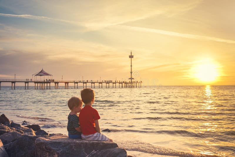 Due ragazzi che si siedono sulla roccia alla spiaggia al tramonto immagine stock libera da diritti Due ragazzi che si siedono sulla roccia alla spiaggia al tramonto immagine stock libera da diritti