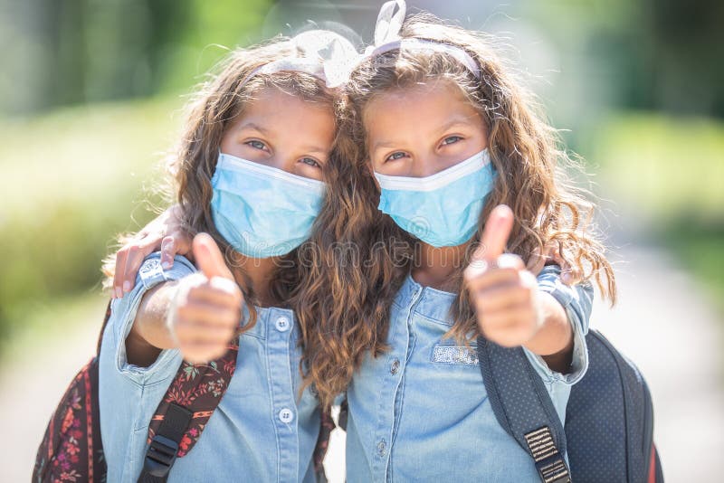 Twin sisters with face masks go back to school during the Covid-19 quarantine and showing thumbs up. Twin sisters with face masks go back to school during the Covid-19 quarantine and showing thumbs up