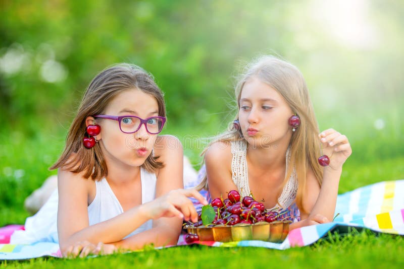 Due sorelle o amici svegli in un giardino di picnic si trovano su una piattaforma e mangiano le ciliege di recente selezionate immagine stock Due sorelle o amici svegli in un giardino di picnic si trovano su una piattaforma e mangiano le ciliege di recente selezionate immagine stock