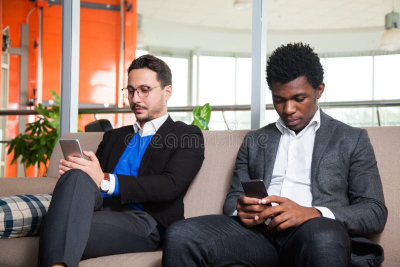 Due uomini multiculturali si siedono sui telefoni cellulari del sofà, di sorriso e della tenuta immagine stock Due uomini multiculturali si siedono sui telefoni cellulari del sofà, di sorriso e della tenuta immagine stock