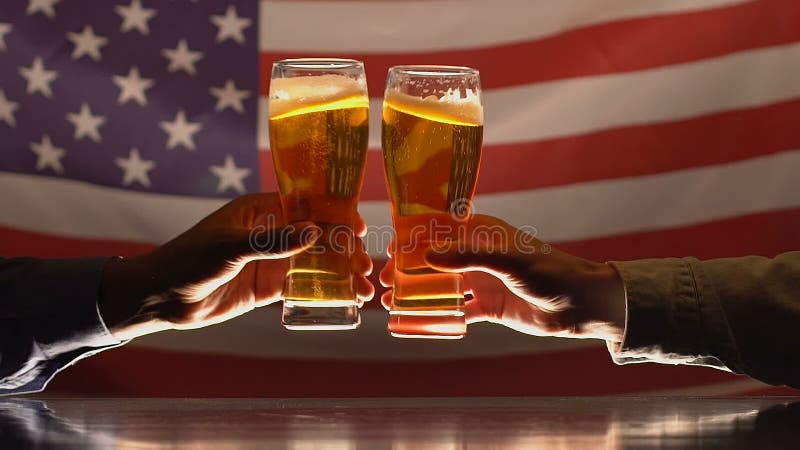 Due vetri di birra tintinnanti degli uomini contro la bandiera di U.S.A., celebrazione di festa dell'indipendenza