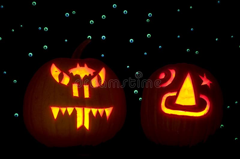 Two carved pumpkins, lighted with candles, are placed in front of a backdrop of hundreds of googly eyes that are lit up with ultraviolet light. One pumpkin is carved as a snake and the other with a sorcerer's hat. Two carved pumpkins, lighted with candles, are placed in front of a backdrop of hundreds of googly eyes that are lit up with ultraviolet light. One pumpkin is carved as a snake and the other with a sorcerer's hat.