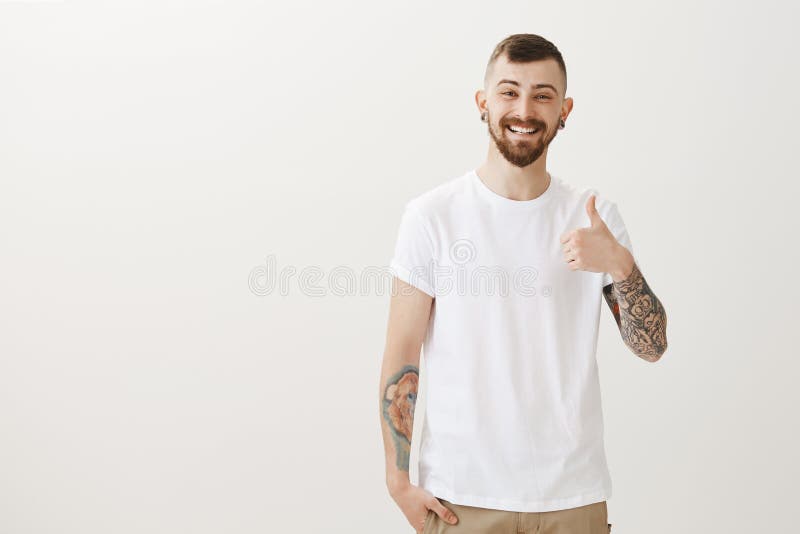 Duimen tonen en portret die van knappe gelukkige mannelijke student met baard en tatoegeringen, het geven cheerfully glimlachen royalty-vrije stock afbeelding Duimen tonen en portret die van knappe gelukkige mannelijke student met baard en tatoegeringen, het geven cheerfully glimlachen royalty-vrije stock afbeelding