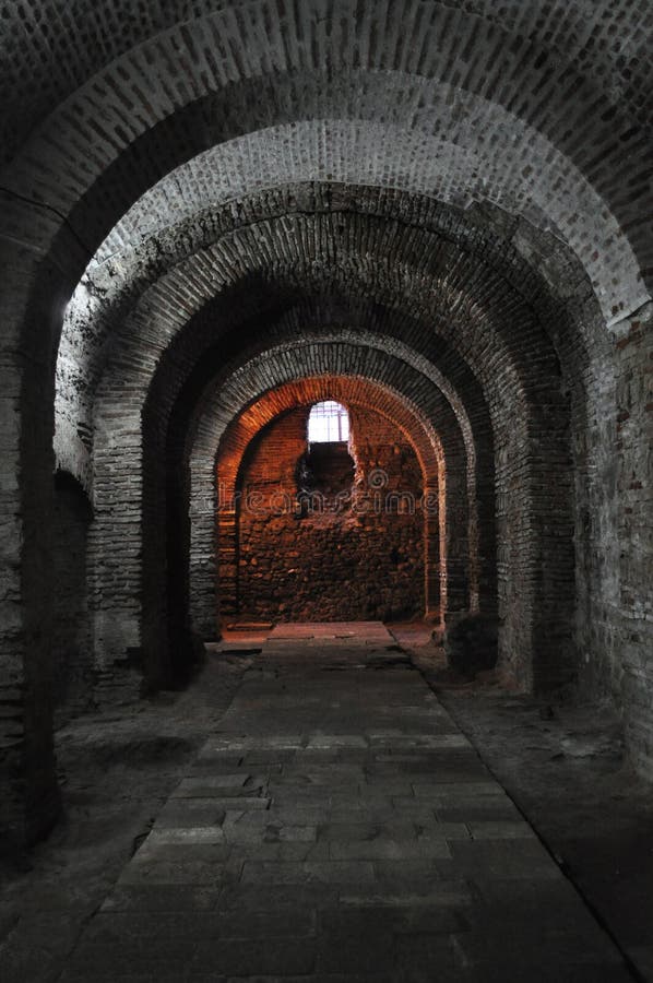 A dark dungeon tunnel with a small window at the far end. The end of the tunnel is red coloured and it has brick walls. The walls are constructed with gothic arcades and made of bricks. A dark dungeon tunnel with a small window at the far end. The end of the tunnel is red coloured and it has brick walls. The walls are constructed with gothic arcades and made of bricks.