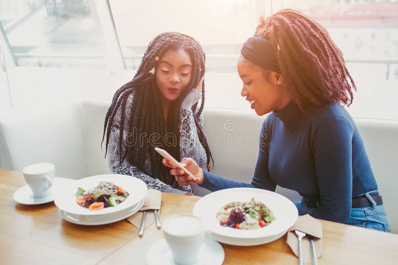 Two african american women sit in cafe at table. They look at phone. Food in balls are on table. Two african american women sit in cafe at table. They look at phone. Food in balls are on table