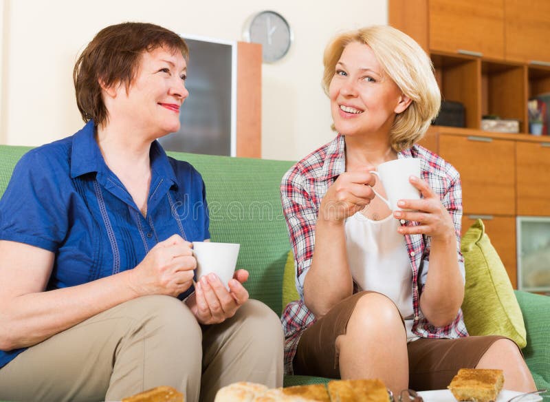 Two smiling mature women friends drinking tea and gossiping at home. Two smiling mature women friends drinking tea and gossiping at home