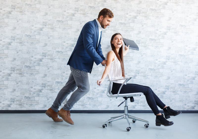 Dwa ffice pracownika ma zabawę przy pracą, facet stacza się dziewczyny na krześle na kołach zdjęcia stock Dwa ffice pracownika ma zabawę przy pracą, facet stacza się dziewczyny na krześle na kołach zdjęcia stock