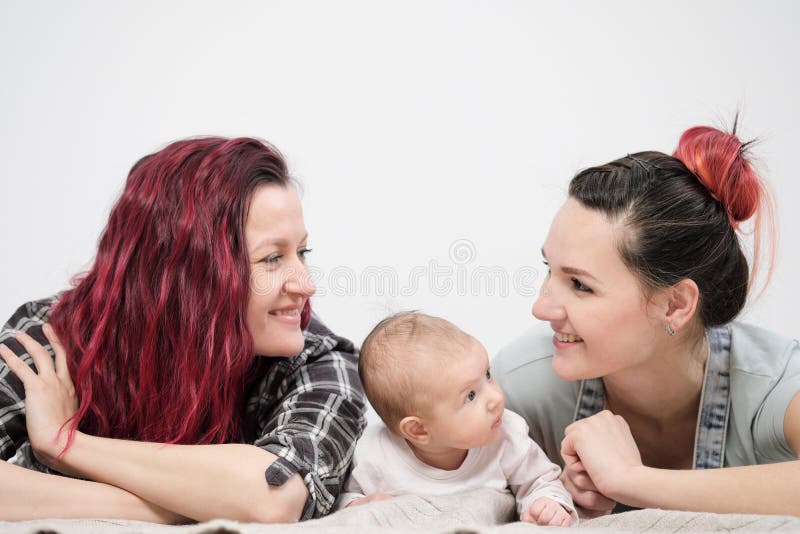 Two young women with dyed red hair and in casual clothes with a baby on a white background. Same-sex marriage and adoption, homosexual lesbian couple. Two young women with dyed red hair and in casual clothes with a baby on a white background. Same-sex marriage and adoption, homosexual lesbian couple.