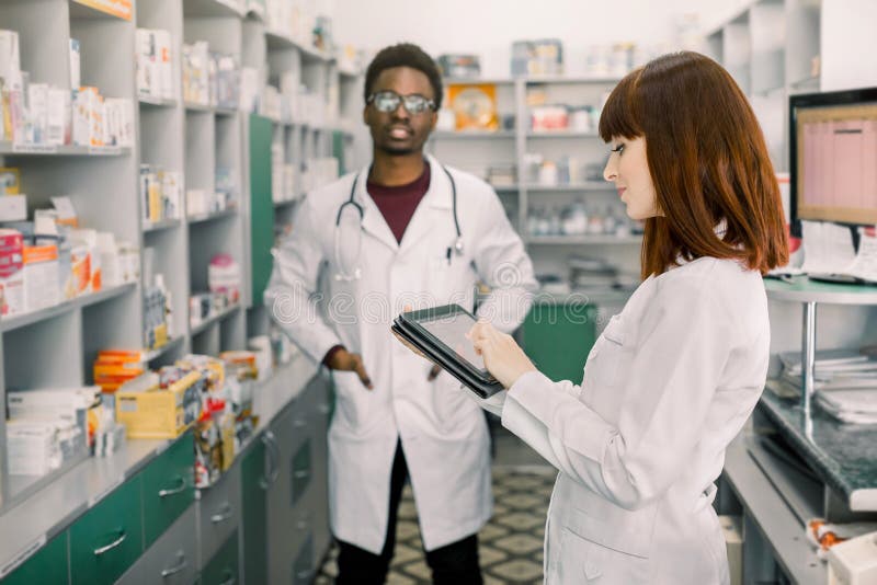 Dwa młodych rozochoconych farmaceut Afrykański mężczyzna i Kaukaska kobieta pracuje wpólnie Kobieta u?ywa pastylk? farmaceuty fotografia stock Dwa młodych rozochoconych farmaceut Afrykański mężczyzna i Kaukaska kobieta pracuje wpólnie Kobieta u?ywa pastylk? farmaceuty fotografia stock