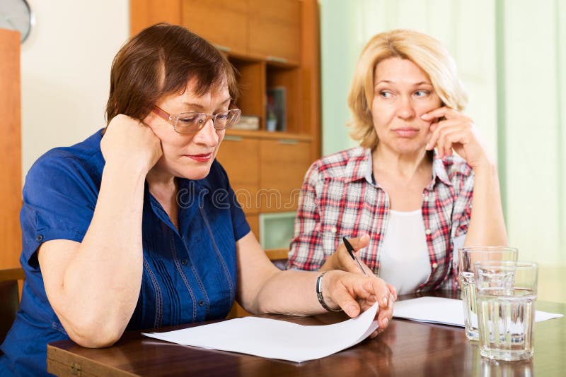 Two sad mature women reading documents at table. Two sad mature women reading documents at table