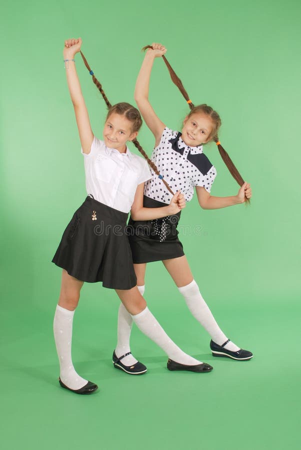 Two school girls plays with plaited hair. Two lovely girls holds plaits of hair in hands and looks in the camera on green. Two school girls plays with plaited hair. Two lovely girls holds plaits of hair in hands and looks in the camera on green