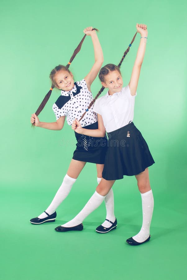 Two school girls plays with plaited hair. Two lovely girls holds plaits of hair in hands and looks in the camera on green. Two school girls plays with plaited hair. Two lovely girls holds plaits of hair in hands and looks in the camera on green