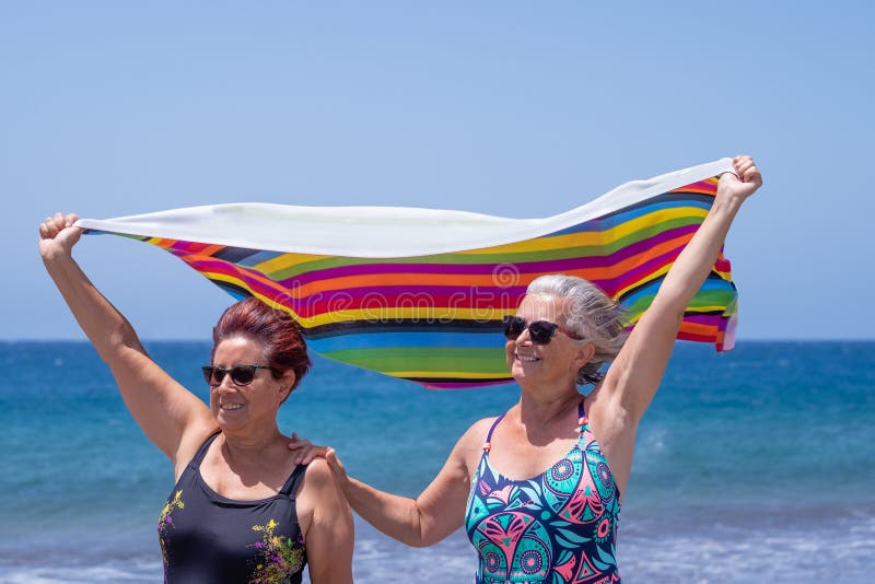 Two mature women enjoying the beach together holding towels in the wind, active retired seniors and vacation concept. Horizon over water. Two mature women enjoying the beach together holding towels in the wind, active retired seniors and vacation concept. Horizon over water.