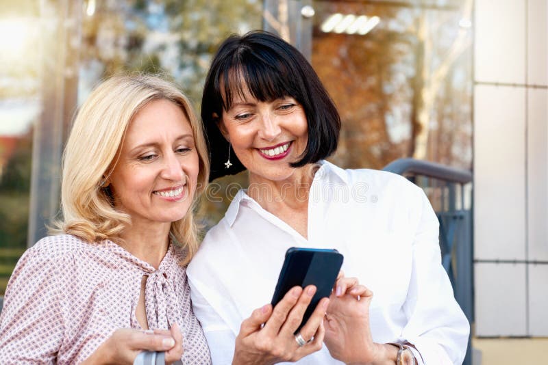 Two mature women looking at the phone and having fun while doing shopping. Pastime concept of mature people. Two mature women looking at the phone and having fun while doing shopping. Pastime concept of mature people