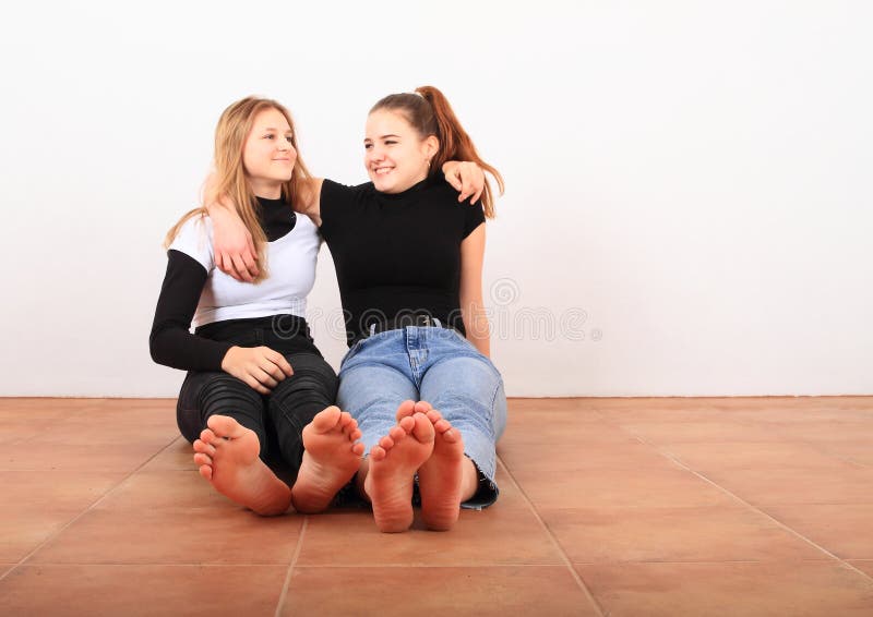 Two smiling teenage girls - friends sitting barefoot on brown tile floor. Friendship concept. Two smiling teenage girls - friends sitting barefoot on brown tile floor. Friendship concept.