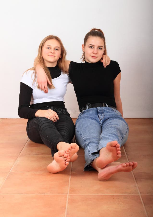 Two smiling teenage girls - friends sitting barefoot on brown tile floor. Friendship concept. Two smiling teenage girls - friends sitting barefoot on brown tile floor. Friendship concept.